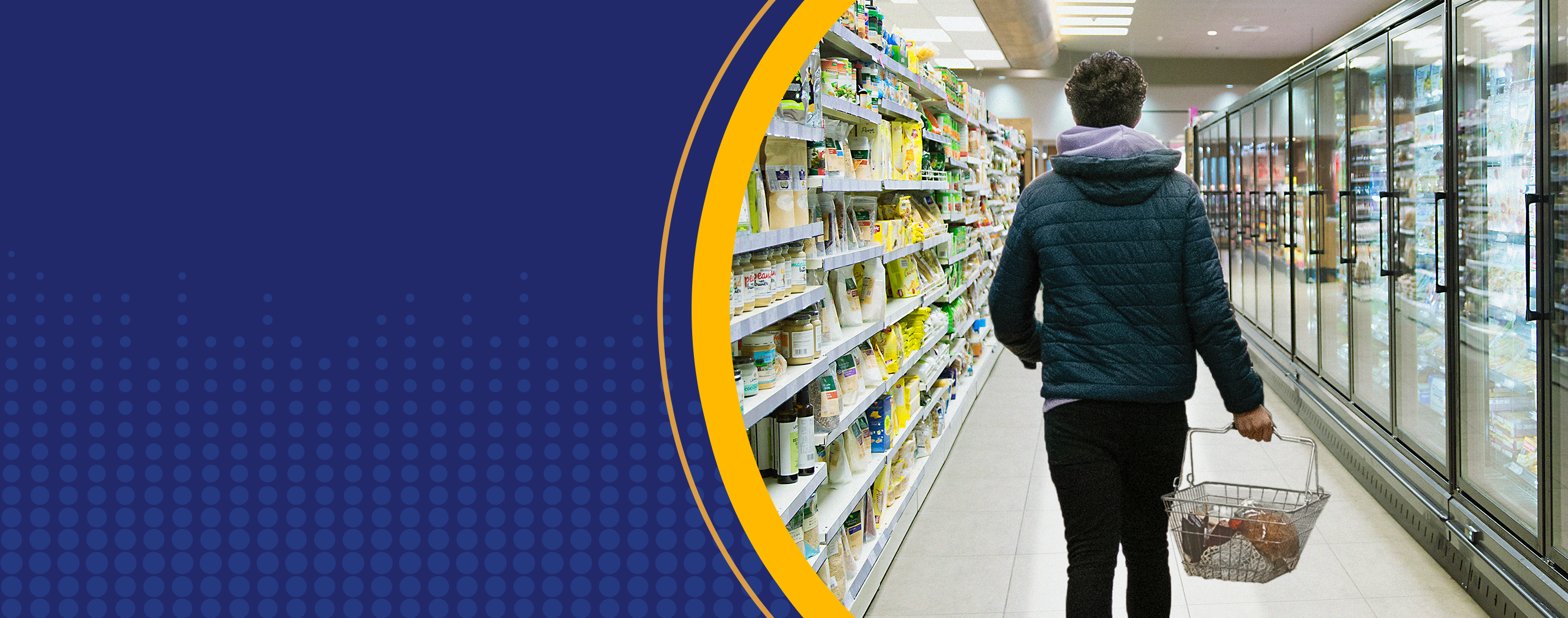 A person in a dark blue, puffy hooded jacket walks away from the camera and down a supermarket aisle with household items stacked on the shelves on both sides. The person carries a shopping basket containing only the few items they could afford as they walk toward the checkout lanes.
